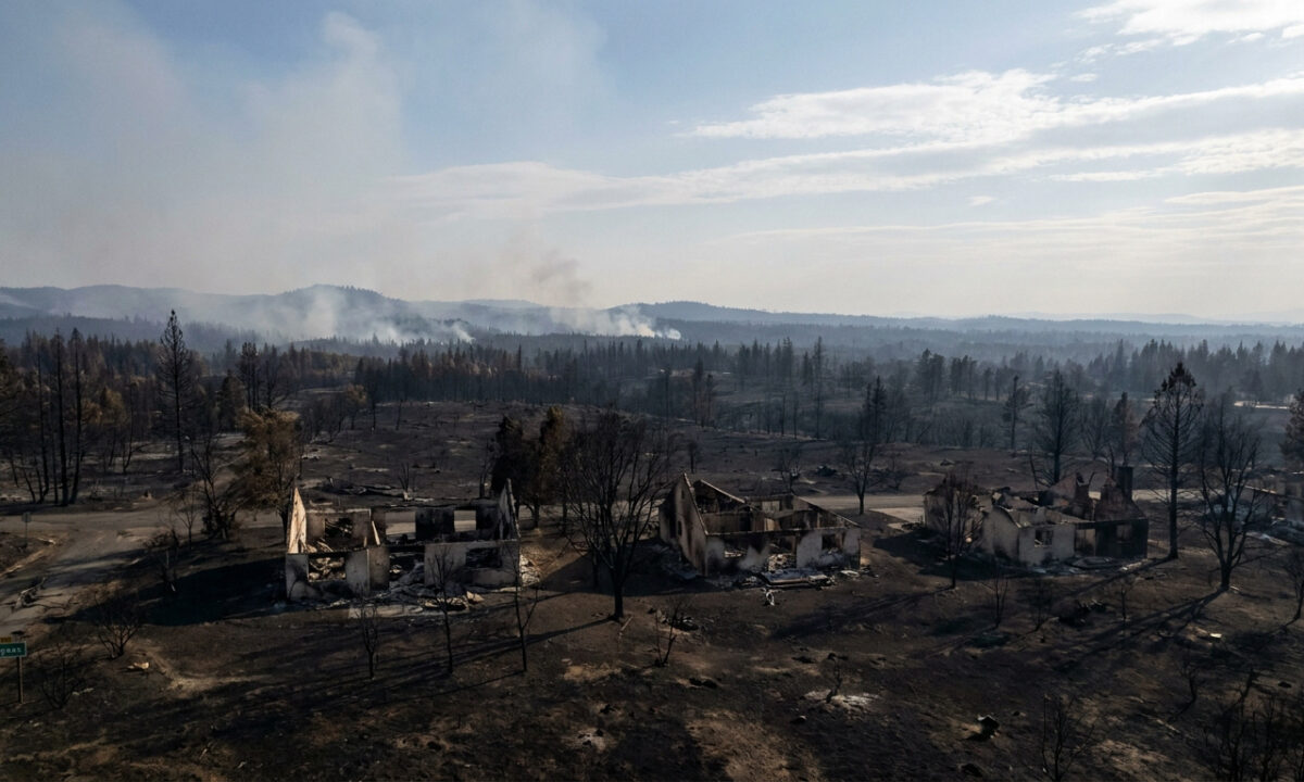 Vue aérienne de maisons détruites par un incendie dans un paysage dévasté par les flammes avec de la fumée à l'horizon.